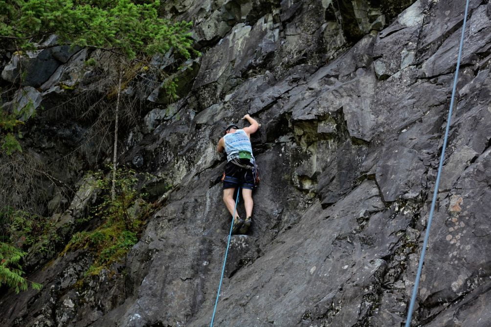 man climbing a dark rock wall with vegetation and ropes